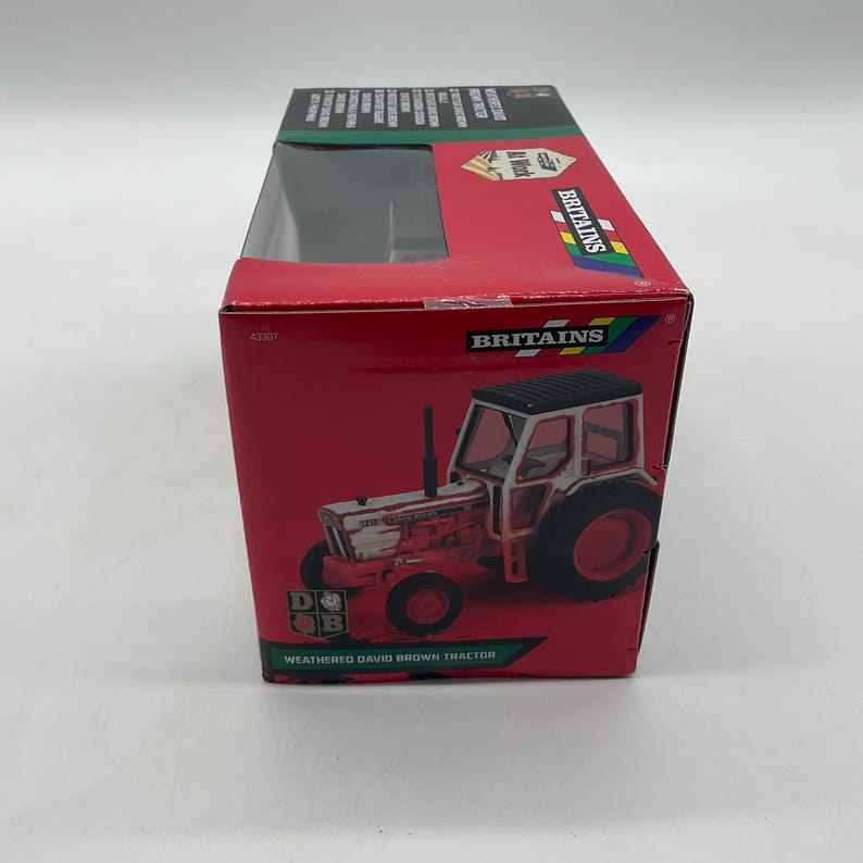 May include: A red box featuring a toy tractor. The tractor is white and red with black tires and a black roof. The box has the word "Britains" and "Weathered David Brown Tractor" printed on it.