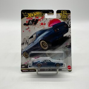 May include: A Hot Wheels die-cast model of a dark blue Nissan Skyline 2000GT-LBWK car. The packaging features Japanese-themed graphics, including a red sun and cherry blossoms. The car is 1/5 in the Japan Historics 5 series.