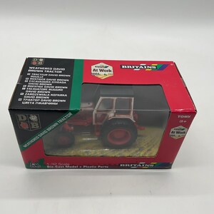 May include: A boxed die-cast model of a weathered David Brown tractor. The tractor is red and white with black tires. The box is red and green with the text "Britains" and "At Work". The scale is 1:32.