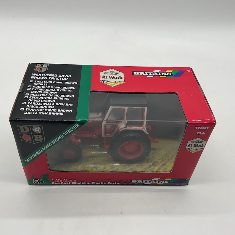 May include: A boxed die-cast model of a weathered David Brown tractor. The tractor is red and white with black tires. The box is red and green with the text "Britains" and "At Work". The scale is 1:32.