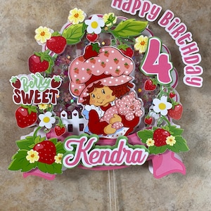 May include: A colorful birthday cake topper featuring a cartoon girl in a strawberry-themed outfit, holding a cat. The topper includes the text "Happy Birthday 4 Kendra" with strawberry and flower accents.