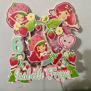 May include: A colorful cake topper featuring Strawberry Shortcake characters. The design includes the number 6, strawberries, flowers, and the name "Isabelle Freya". The topper has a glittery finish and is designed for a birthday celebration.