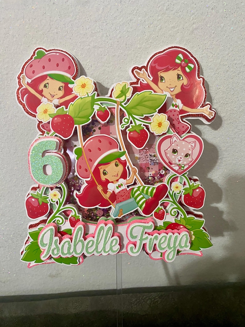 May include: A colorful cake topper featuring Strawberry Shortcake characters. The design includes the number 6, strawberries, flowers, and the name "Isabelle Freya". The topper has a glittery finish and is designed for a birthday celebration.