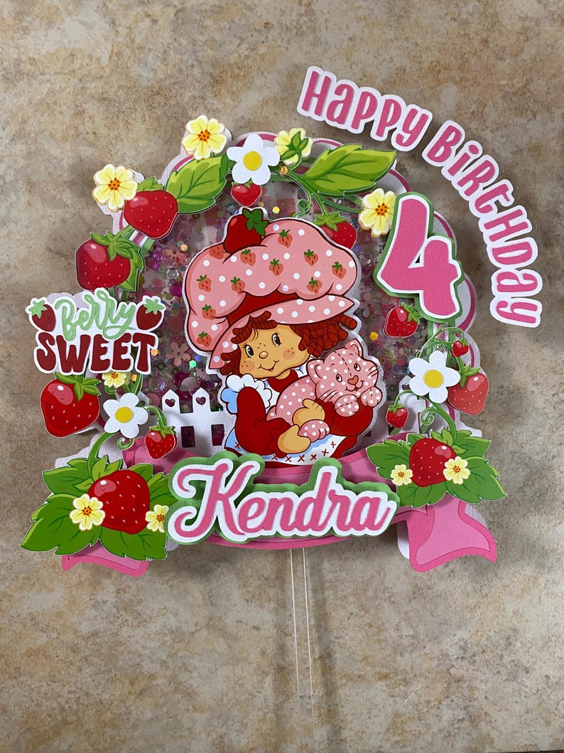 May include: A colorful birthday cake topper featuring a cartoon girl in a strawberry-themed outfit, holding a cat. The topper includes the text "Happy Birthday 4 Kendra" with strawberry and flower accents.
