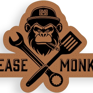 Può includere: Un cerotto in pelle marrone con una silhouette nera di una scimmia che indossa un berretto da baseball e tiene una chiave inglese e una spatola. Il testo "GREASE MONKEY" è stampato in nero sul cerotto.