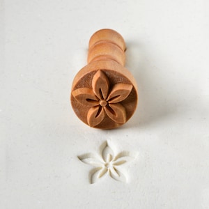 May include: A wooden stamp with a flower design is used to create an impression in white clay. The stamp has a wooden handle and a round base with a carved flower design.