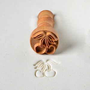May include: A wooden cherry-shaped cookie stamp with a handle. The stamp is used to create an impression of two cherries with a stem and leaves in dough.
