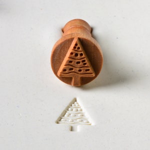May include: A wooden Christmas tree stamp with a detailed design. The stamp is used to create an impression of a Christmas tree on a surface.