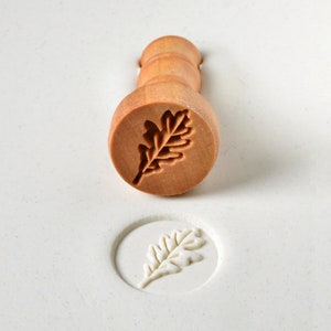 May include: A wooden stamp with an oak leaf design. The stamp is used to create an impression in clay or dough.