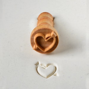 May include: A wooden heart-shaped stamp with an arrow through it. The stamp is used to create an impression in clay or dough.