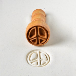 May include: A wooden peace sign stamp with a brown handle. The stamp has been used to create a white peace sign impression on a white surface.