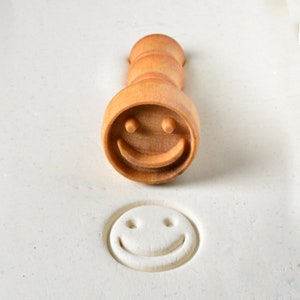 May include: A wooden smiley face cookie cutter with a handle. The cutter has been used to create a smiley face impression in white dough.