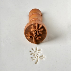 May include: A wooden stamp with a floral design. The stamp is used to create an impression of a flower in clay or dough.