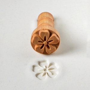 May include: A wooden flower-shaped stamp with a handle. The stamp is used to create an impression of a flower in clay or dough.