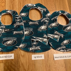 May include: Three baby bibs with a Philadelphia Eagles theme. The bibs are green with white and black accents. The bibs have different sizes and styles. The text on the bibs reads "Philadelphia Eagles".