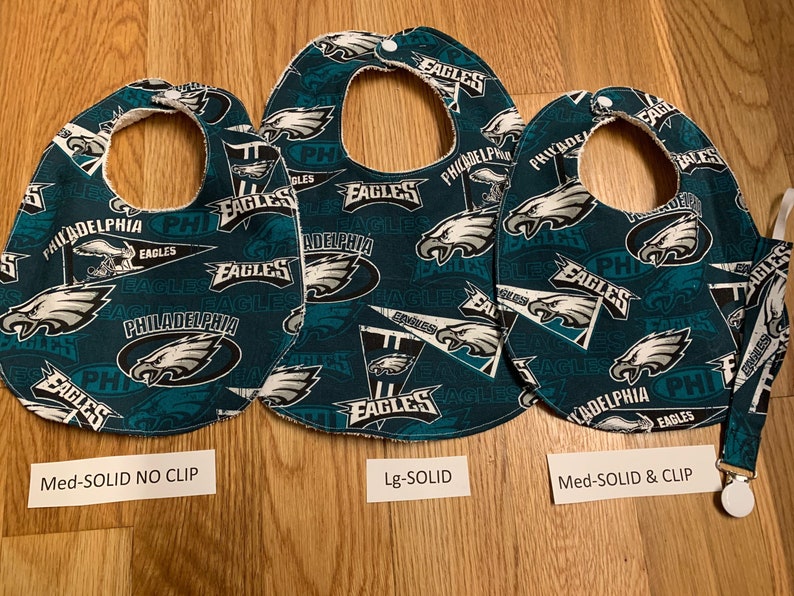 May include: Three baby bibs with a Philadelphia Eagles theme. The bibs are green with white and black accents. The bibs have different sizes and styles. The text on the bibs reads "Philadelphia Eagles".