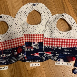 May include: Three baby bibs with a red and white checkered pattern and a blue and white Patriots themed fabric. The bibs are labeled "Med. Mix", "Lg. Mix", and "Med. Mix + Clip".