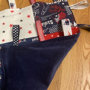 May include: A blue and white baby blanket with various fabric textures and ribbons. The blanket features a red, white, and blue American flag pattern, a red star pattern, and a blue pattern with white stars. The blanket also has the words "Patriots" and "Football" printed on it.