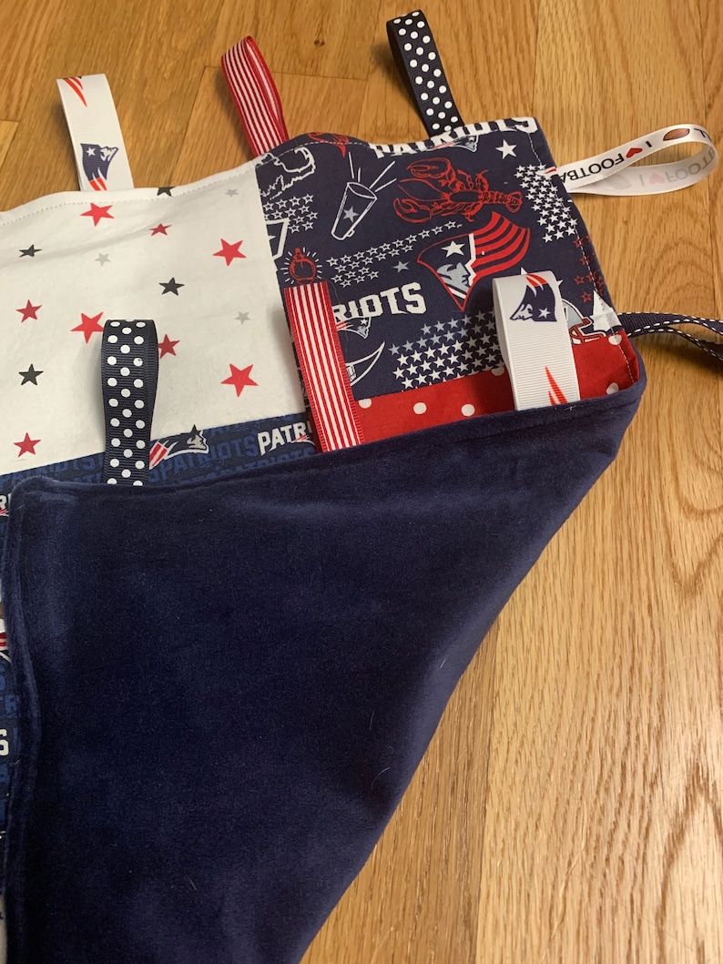 May include: A blue and white baby blanket with various fabric textures and ribbons. The blanket features a red, white, and blue American flag pattern, a red star pattern, and a blue pattern with white stars. The blanket also has the words "Patriots" and "Football" printed on it.
