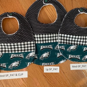 May include: Three baby bibs with a black and white polka dot pattern, a black and white checkered pattern, and a green pattern with the word "Eagles" repeated. The bibs are labeled "Med-SP_PAT & CLIP", "Lg-SP_PAT", and "Med-SP_PAT NO CLIP".