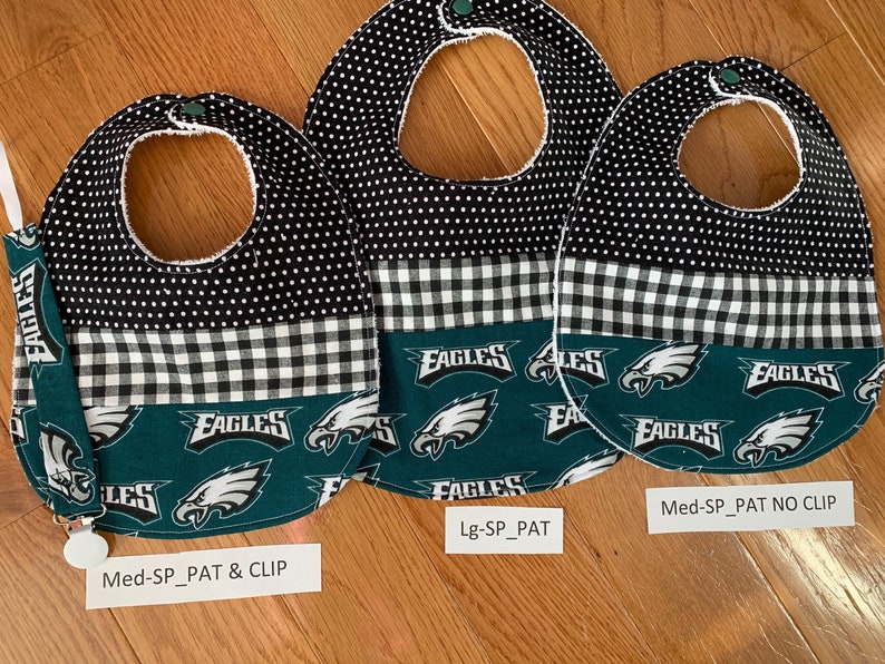 May include: Three baby bibs with a black and white polka dot pattern, a black and white checkered pattern, and a green pattern with the word "Eagles" repeated. The bibs are labeled "Med-SP_PAT & CLIP", "Lg-SP_PAT", and "Med-SP_PAT NO CLIP".