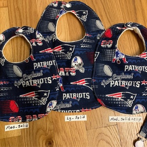 May include: Three baby bibs with a New England Patriots theme. The bibs are blue with red, white, and silver accents. The bibs have a variety of Patriots logos and text, including "New England Patriots" and "Patriots". The bibs are available in three sizes: medium, large, and extra large. The medium bib has a solid back, the large bib has a solid back, and the extra large bib has a solid back with a clip.