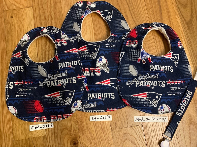 May include: Three baby bibs with a New England Patriots theme. The bibs are blue with red, white, and silver accents. The bibs have a variety of Patriots logos and text, including "New England Patriots" and "Patriots". The bibs are available in three sizes: medium, large, and extra large. The medium bib has a solid back, the large bib has a solid back, and the extra large bib has a solid back with a clip.