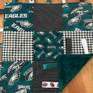 May include: A green, black, and white baby blanket with various textures and patterns. The blanket features the Philadelphia Eagles logo and the words "Philadelphia Eagles". The blanket has several fabric loops and tags for a baby to play with.