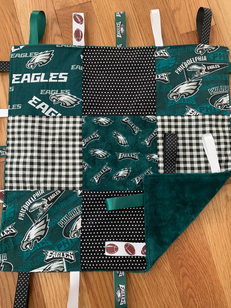 May include: A green, black, and white baby blanket with various textures and patterns. The blanket features the Philadelphia Eagles logo and the words "Philadelphia Eagles". The blanket has several fabric loops and tags for a baby to play with.