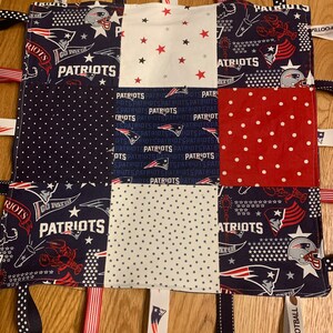 May include: A blue and white baby blanket with a patchwork design featuring the New England Patriots logo and the words "Patriots" and "Go Patriots". The blanket has several fabric tags attached to the edges, including a tag that says "I Love Football".