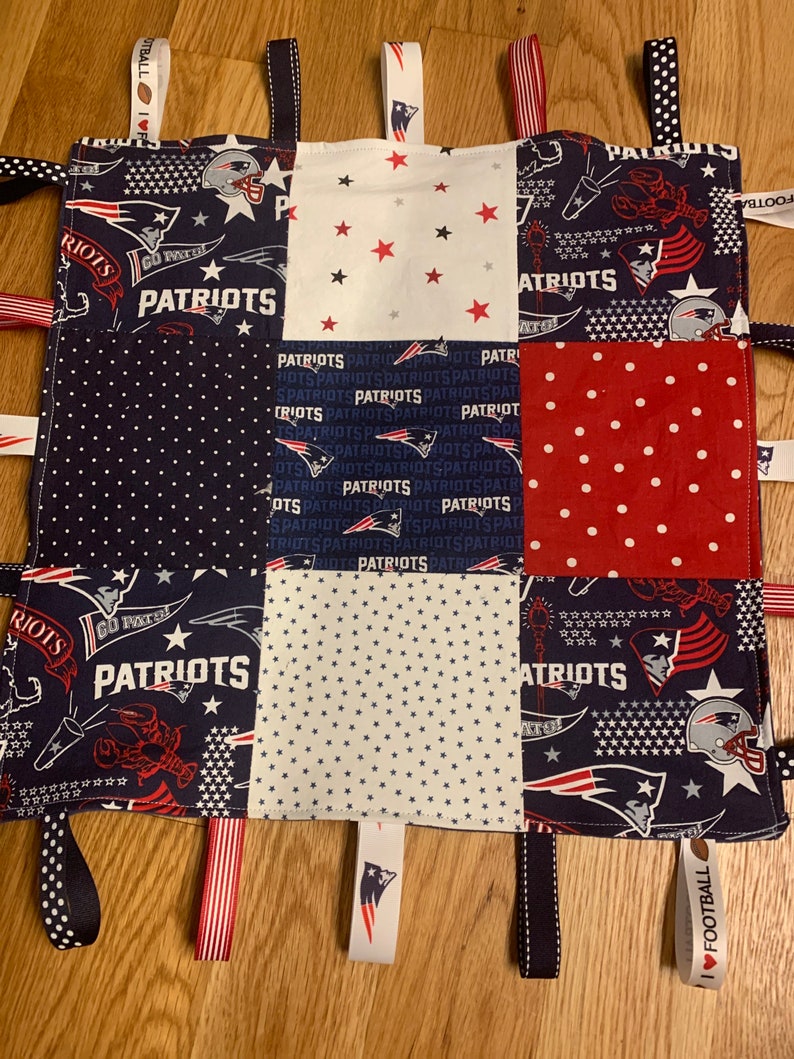 May include: A blue and white baby blanket with a patchwork design featuring the New England Patriots logo and the words "Patriots" and "Go Patriots". The blanket has several fabric tags attached to the edges, including a tag that says "I Love Football".