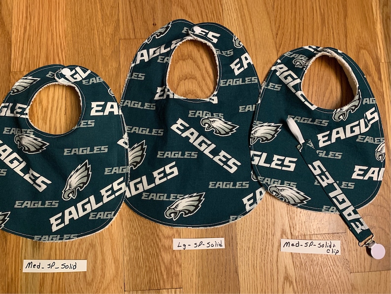 May include: Three green and white baby bibs with a Philadelphia Eagles pattern. The bibs are labeled "Med - SP - Solid", "Lg - SP - Solid", and "Med - SP - Solid + clip".