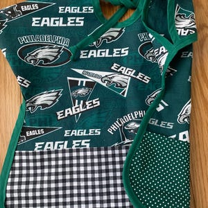May include: Green and white fabric apron with a pocket. The apron has a checkered pattern on the bottom and a green fabric with white polka dots on the pocket. The apron is decorated with the Philadelphia Eagles logo and the words "Eagles" repeated throughout.