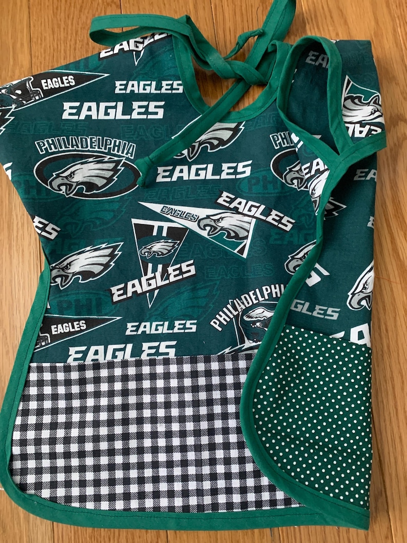 May include: Green and white fabric apron with a pocket. The apron has a checkered pattern on the bottom and a green fabric with white polka dots on the pocket. The apron is decorated with the Philadelphia Eagles logo and the words "Eagles" repeated throughout.