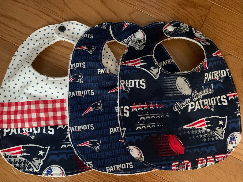 May include: Three blue and white fabric baby bibs with a New England Patriots theme. The bibs feature a red, white, and blue checkered pattern, stars, and the team logo.