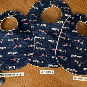 May include: Three blue and white baby bibs with a Patriots logo print. The bibs are made of soft fabric and have a white trim. The bibs are labeled with different sizes: Med-Mini Print & Clip, Lg-Mini Print, and Med-Mini Print.