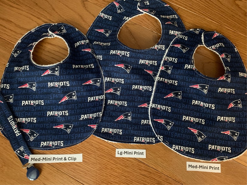 May include: Three blue and white baby bibs with a Patriots logo print. The bibs are made of soft fabric and have a white trim. The bibs are labeled with different sizes: Med-Mini Print & Clip, Lg-Mini Print, and Med-Mini Print.