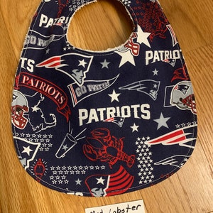 May include: A blue and red baby bib with a New England Patriots theme. The bib features a variety of Patriots logos, including the team's logo, the words "Go Pats!" and "Patriots", and a lobster. The bib also features stars and the words "Med. Lobster".