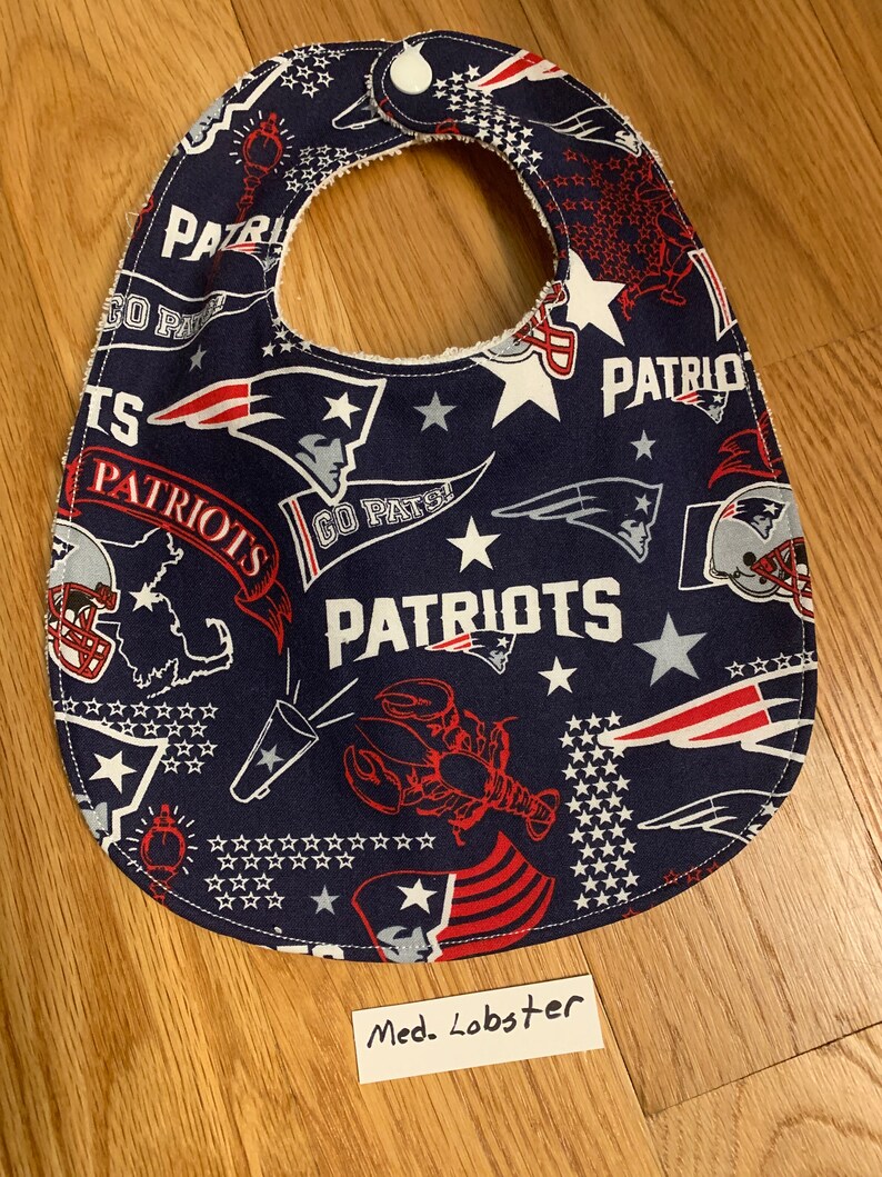 May include: A blue and red baby bib with a New England Patriots theme. The bib features a variety of Patriots logos, including the team's logo, the words "Go Pats!" and "Patriots", and a lobster. The bib also features stars and the words "Med. Lobster".