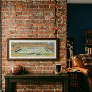 May include: A rectangular framed panoramic print depicting a football stadium filled with spectators, featuring a green field with yard lines and a central logo, displayed in a dark wood frame on a rustic red brick wall above a wooden console table.