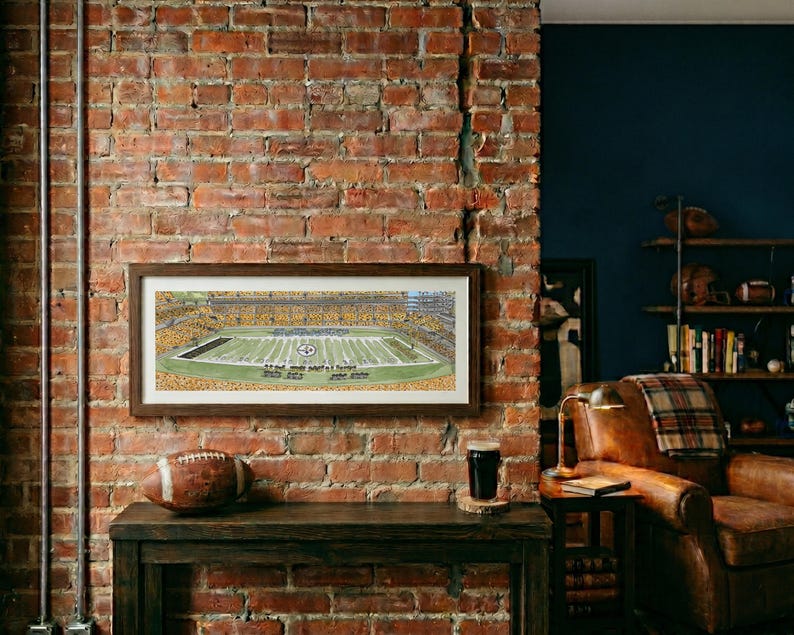 May include: A rectangular framed panoramic print depicting a football stadium filled with spectators, featuring a green field with yard lines and a central logo, displayed in a dark wood frame on a rustic red brick wall above a wooden console table.