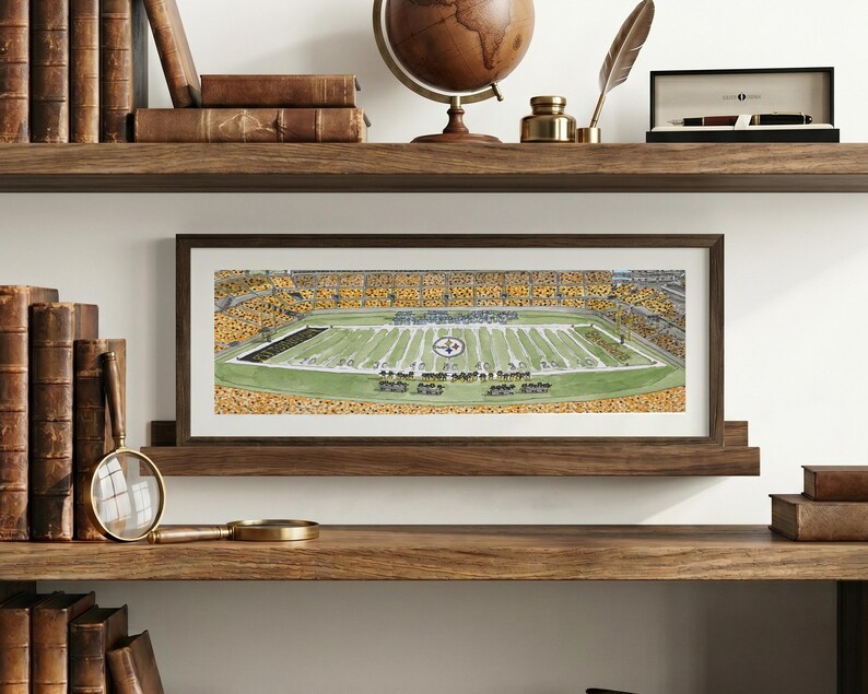 May include: A rectangular framed print featuring a detailed illustration of a football stadium with a green field, yard lines, and a crowd of spectators in yellow and black. The artwork is displayed in a dark wood frame on a wooden shelf.