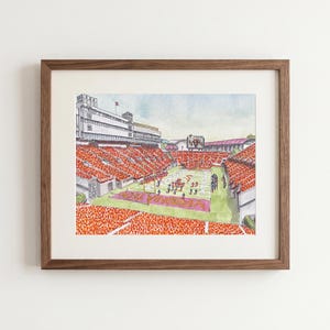 May include: A framed watercolor print depicting a football stadium filled with spectators wearing orange. The field features the text Virginia Tech in the end zone, with players positioned on the green turf under a light blue sky.
