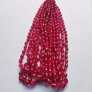 May include: A collection of numerous translucent, deep pink, teardrop-shaped beads arranged in multiple vertical strands. Each bead features a smooth, polished surface that catches the light, creating a uniform and vibrant appearance throughout the set.