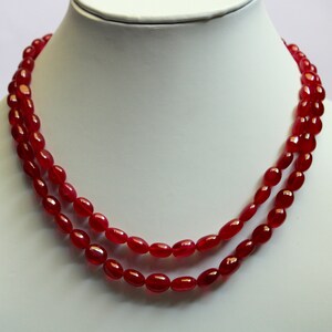 May include: A double-strand necklace featuring smooth, polished, oval-shaped red beads. The beads are uniform in size and color, creating a continuous, vibrant red loop that rests against the white display form, showcasing a classic design.