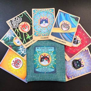 May include: A deck of My Furry Mystic Cat Cards arranged in a fan shape around a teal fabric pouch. Each rectangular card features a unique cat illustration in a circular frame with a small heart above the head and various colourful backgrounds.