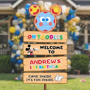 Pode incluir: Uma placa de madeira vertical com um topo de Toodles e quatro tábuas. As tábuas exibem o texto: OH TOODLES, WELCOME TO com um gráfico do Mickey Mouse, ANDREW'S 1ST BIRTHDAY e COME INSIDE IT'S FUN INSIDE com um ícone de luva branca.