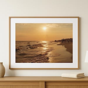 May include: A framed photograph of a beach scene at sunset. The sun is a bright orb in the sky, casting a golden glow over the water and sand. Silhouetted figures walk along the shoreline.