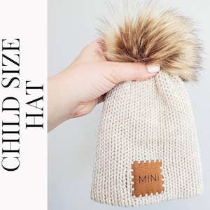 May include: A cream-colored knitted hat with a brown faux fur pom pom and a brown leather patch that says "MINI".