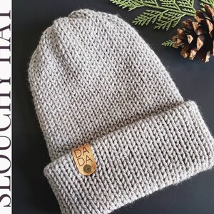 May include: A gray knit slouchy hat with a folded brim. A small leather tag with the text "DA DA" is attached to the brim. The hat is made of a thick, textured yarn.